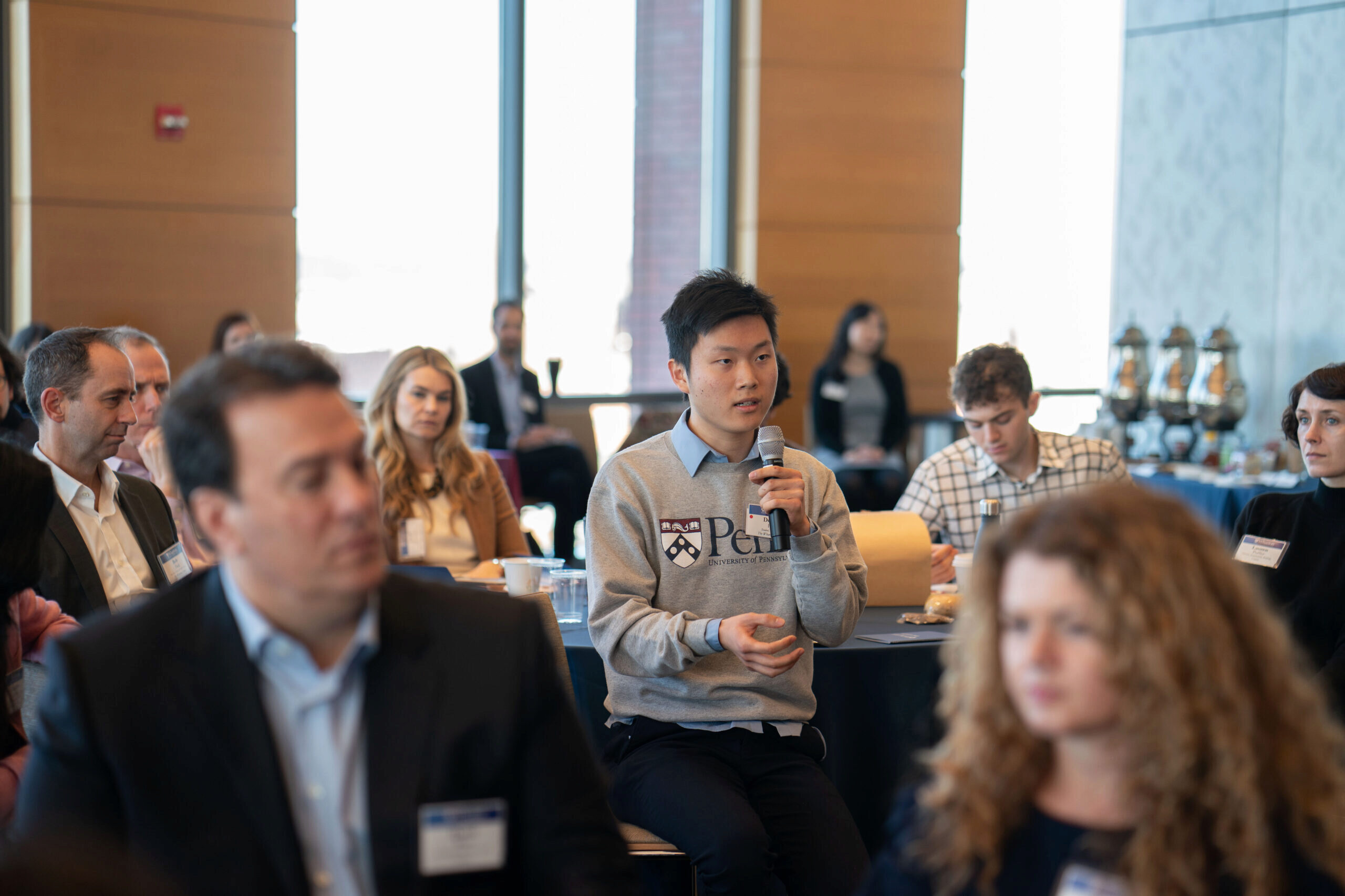 Student in a Penn crew neck and button down shirt holding a microphone and reporting back his group's discussion